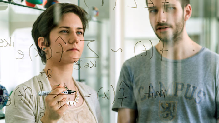 A woman and a man look at a scientific formula written on a glass board.