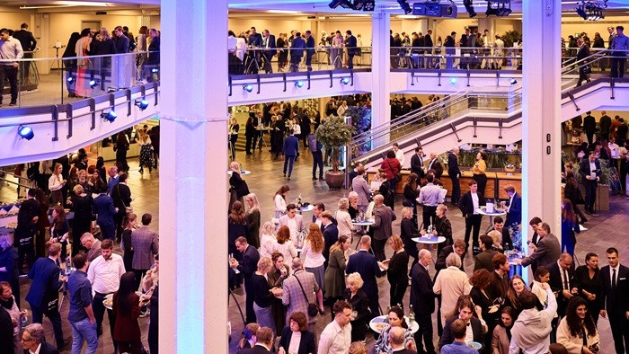 Große Halle mit vielen Gästen an Stehtischen auf zwei Ebenen, lebhafte Gespräche und festliche Beleuchtung. 