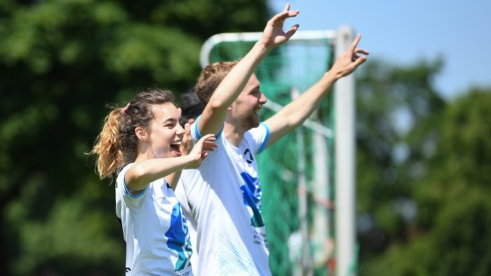 A young woman and a young man in soccer jerseys cheer on the sidelines.