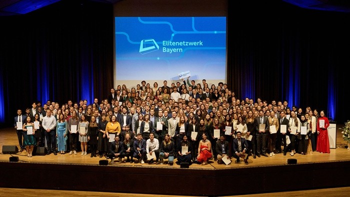 © Florian Freund Großes Gruppenfoto auf der Bühne vor dem Logo „Elitenetzwerk Bayern“; zahlreiche Absolventinnen und Absolventen mit Urkunden.