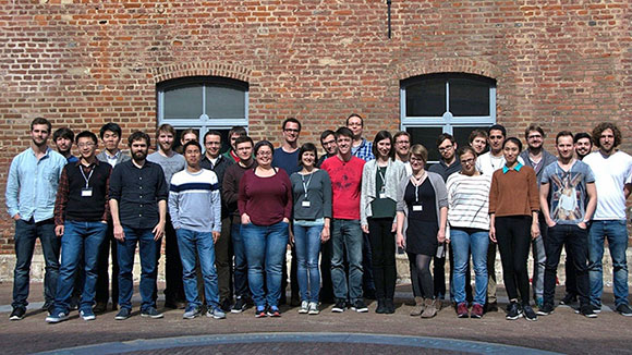 © IDK Topologische Isolatoren Eine Gruppe von Personen steht an einem warmen Tag vor einem Backsteingebäude zum Gruppenfoto aufgestellt und blickt in die Kamera.
