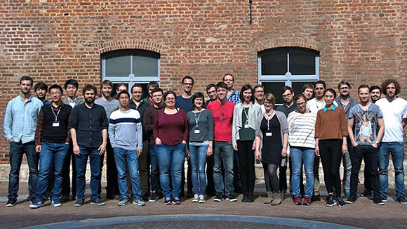 A group of people is standing on a warm day in front of a brick building to the group photo and looks into the camera. 