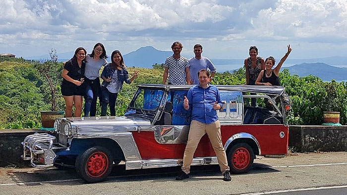 Group picture: Young people, positioned around an old car, laugh at the camera. In the background are green hills, mountains and blue sky.