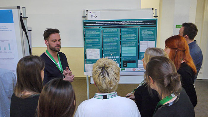 A speaker in front of an audience presenting a poster visible in the background.