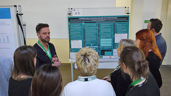 A speaker in front of an audience presenting a poster visible in the background. 