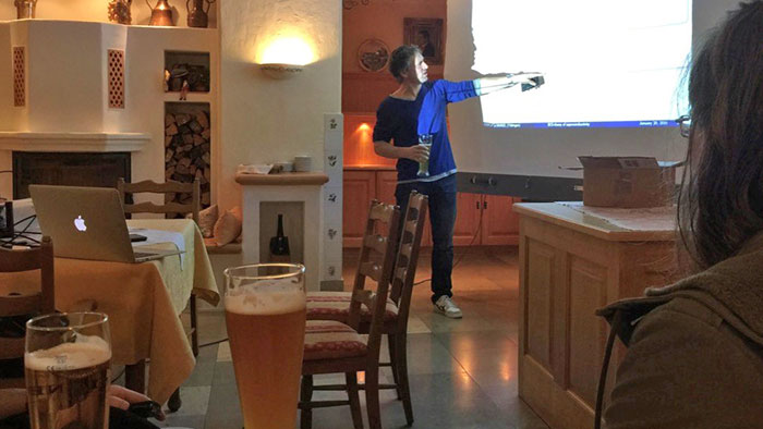A man standing in front of a screen pointing to it.  He is giving a talk holding a beer glass in his other hand. In the foreground, there are more beer glasses.