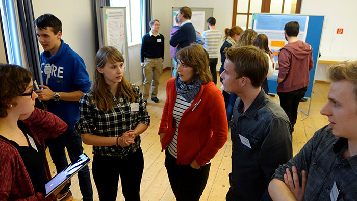 Students standing in a half-circle and talking. In the background you can see other persons looking at posters and talking about them.