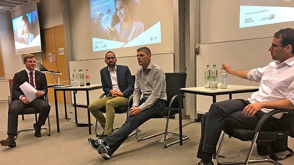 Four people sit next to each other on chairs and discuss. In the background, a presentation on canvases is seen. 