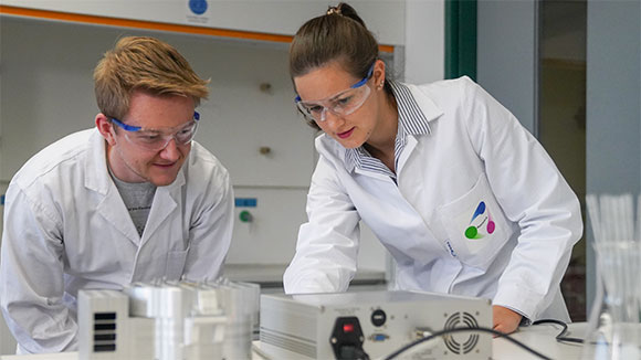 A woman and a man dressed in lab coats and wearing protective goggles look at a box-shaped photo reactor standing on a table. The woman is adjusting something on the device.