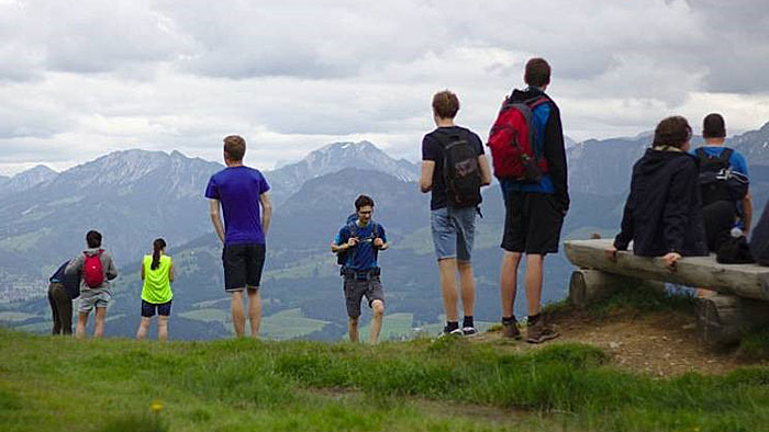 A group of young people, all dressed for a moderate hiking trip, reaching the top of a hill on a cloudy day. Some are looking to a mountain chain in the distance.