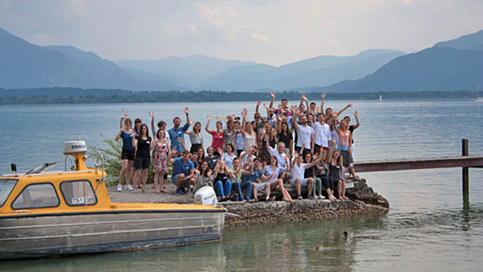 Young people gathered for a group photo on a jetty and waving into the camera. Behind them is a lake, surrounded by the Alps.