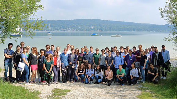 Gruppenbild: Eine Vielzahl von Menschen stehen zu einem Gruppenfoto aufgestellt und von Bäumen umrandet am Ufer eines Sees.