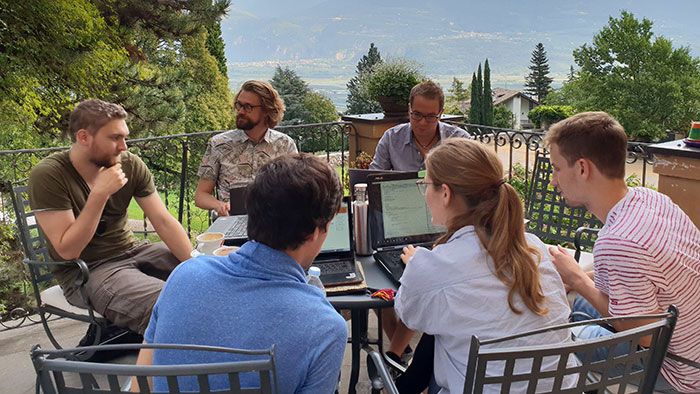 Junge Menschen sitzen auf einer Terrasse im Gespräch vertieft um einen Tisch vor einem Bergpanorama.