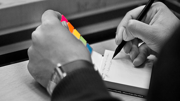 © Matthias Deininger Detailaufnahme: Die linke Hand hält einen Haftnotizblock aufgeklappt, während die rechte Hand einen Bleistift hält und etwas auf ein Post-It schreibt.