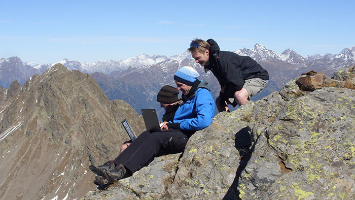 Two young men sit on a rock on top of a mountain, a third student cowers behind them and looks over their shoulders. The two sitting students wear caps and focus on their notebooks. 