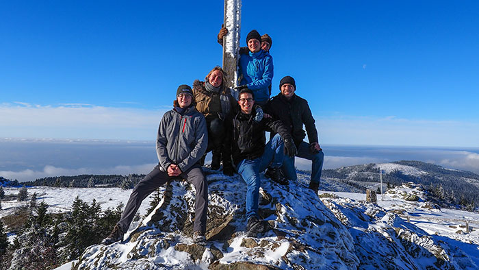 Students in winter clothes around a summit cross and smile into the camera.