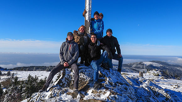 Gruppenbild: Studierende stellen sich in Winterkleidung um ein Gipfelkreuz und lächeln in die Kamera.