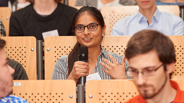 A young woman sits among other attendees in a lecture hall and speaks into a microphone.