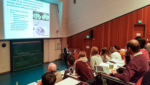 The picture shows a fully occupied lecture hall. The lecturer stands behind a lectern in front of a screen showing a presentation slide with immune-histochemical pictures. 