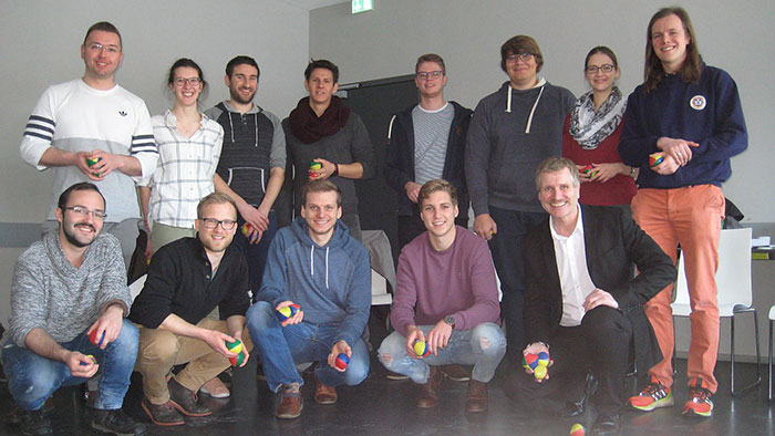 Gruppenbild: Eine Gruppe junger Studierender sowie der Seminarleiter sind in einem Seminarraum zu einem Gruppenfoto aufgestellt. Jonglierbälle liegen vor Ihnen auf dem Boden, manche der Studierenden halten Jonglierbälle in ihren Händen.