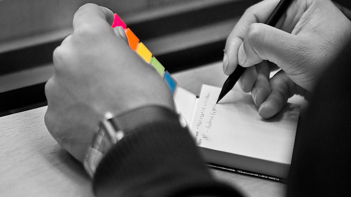 Detail image: A person‘s left hand is holding an opened notebook, while the right hand is taking a note with a pencil.