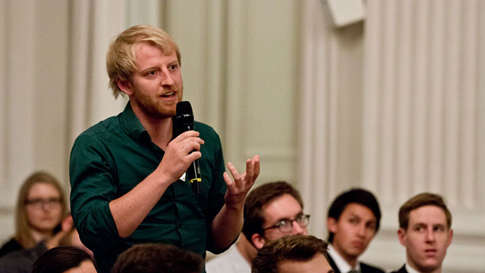 A young man stands in the middle of sitting people and animatedly speaks into a microphone. 