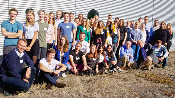 Students and lecturers in front of a grey building on a meadow to take a group photo 