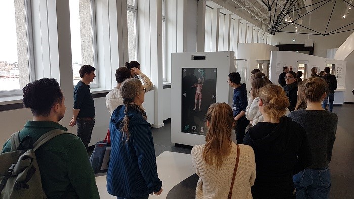 Eine Gruppe steht vor einem Bildschirm, auf dem ein Muskelmodell als Spiegelbild eines Teilnehmers zu sehen ist. 