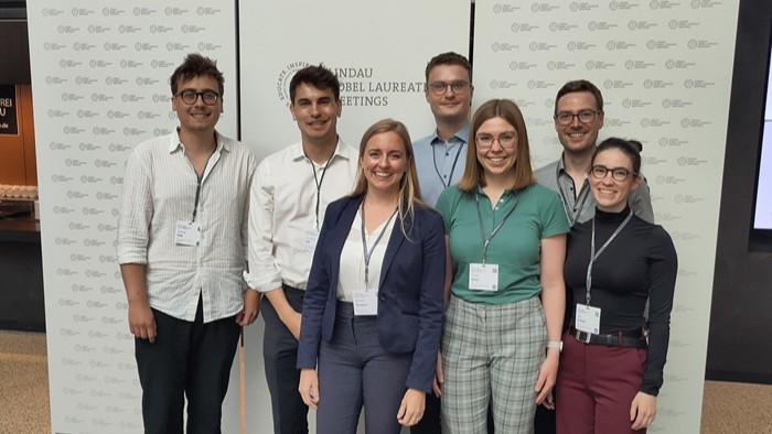Seven young participants stand smiling in front of a photo wall of the “Lindau Nobel Laureate Meetings” and convey an impression of community and scientific commitment.
