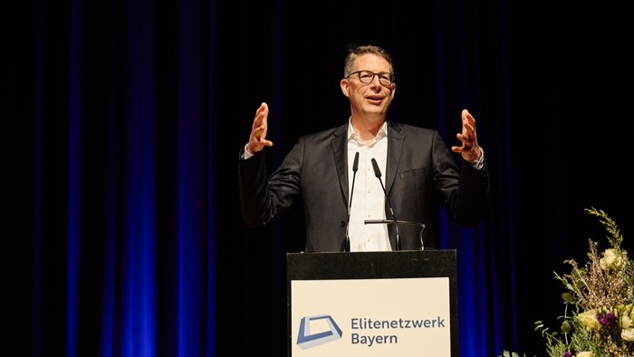 Eine Person spricht am Rednerpult mit dem Logo „Elitenetzwerk Bayern“, im Hintergrund dunkler Vorhang und Blumendekoration. 