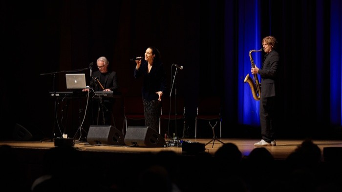 Drei Musiker auf einer Bühne: links Keyboard mit Laptop, in der Mitte eine Person am Mikrofon, rechts eine Person mit Saxofon, vor blauem Bühnenlicht. 