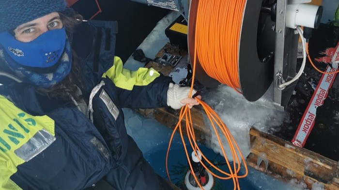 Das Bild zeigt eine Person in Winterbekleidung mit Gesichtsmaske, die ein Bündel orangefarbener Kabel hält. Daran hängt eine Sonde, die in ein eisiges Gewässer eingetaucht wird.