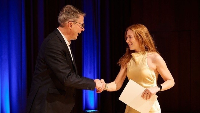 Zwei Personen auf einer Bühne beim Handschlag; eine Person hält eine Urkunde, im Hintergrund blaues Licht und Vorhang. 