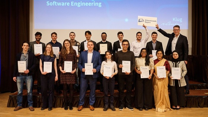 Eine Gruppe steht auf der Bühne in zwei Reihen, viele halten Urkunden; im Hintergrund die Projektionsfläche mit Programmtitel. 