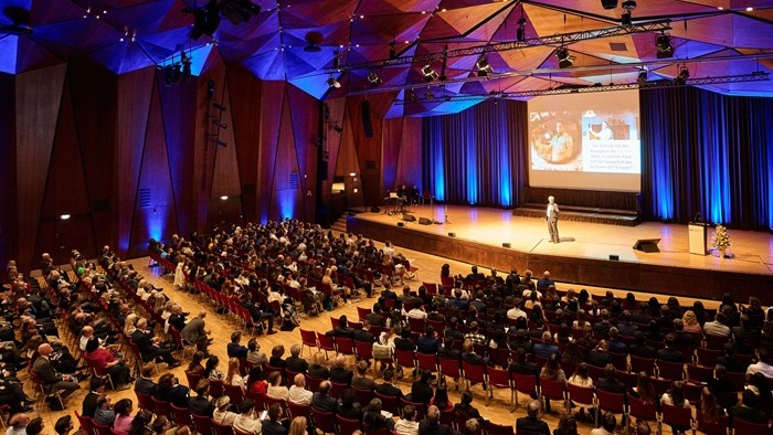 Ein großer Saal mit vielen Sitzreihen; vorne eine Person auf der Bühne vor Leinwand, das Publikum hört aufmerksam zu. 