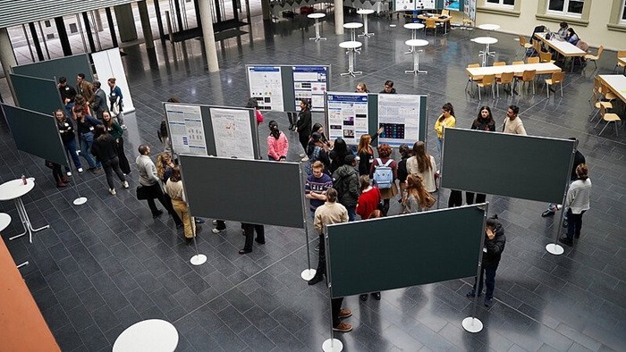 Menschen betrachten wissenschaftliche Poster auf einer Ausstellung in einer großen Halle mit Tischen und Stühlen im Hintergrund.