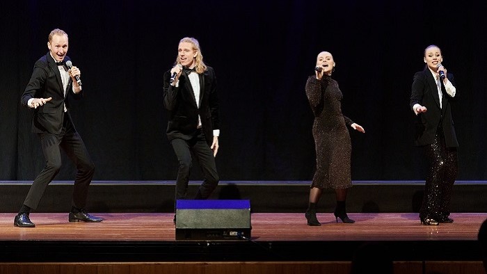 Two young women and two young men in festive black dresses are singing and dancing on stage.