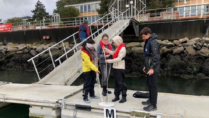 Three people stand on a footbridge and pull up a small container with a narrow rope. 