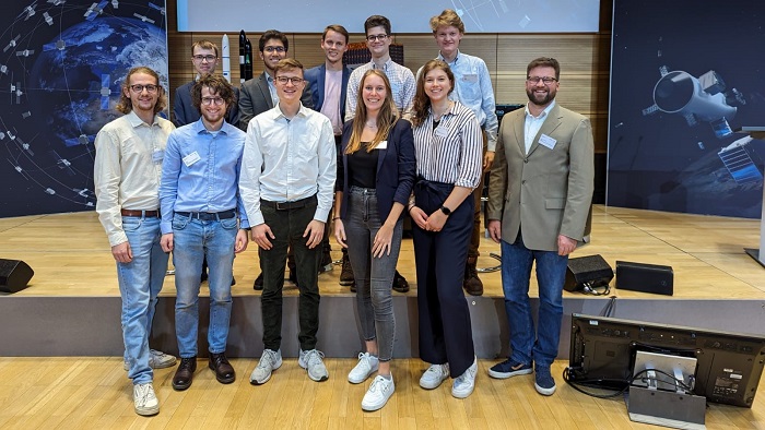 Die Studierenden des Elitestudiengangs „Satellite Technology“ machen ein Gruppenfoto gemeinsam mit Prof. Dr. Guido Dietl vor der Bühne der Kleinsatellitenkonferenz 2023.