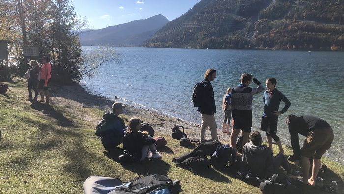 Studierende sitzen am Ufer des Walchensees, einige mit nackten Füßen.