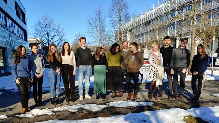 A group of 13 doctoral students from the IDK and the Research Group are standing next to a building. Some of the doctoral students are looking at each other, others are looking at the camera.