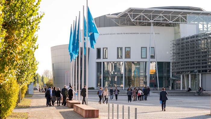 The image shows a group of people walking toward a modern building with large windows and several blue flags on flagpoles in front of it.