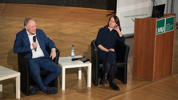 A man talks into a microphone at a panel discussion while a woman next to him looks at him.
