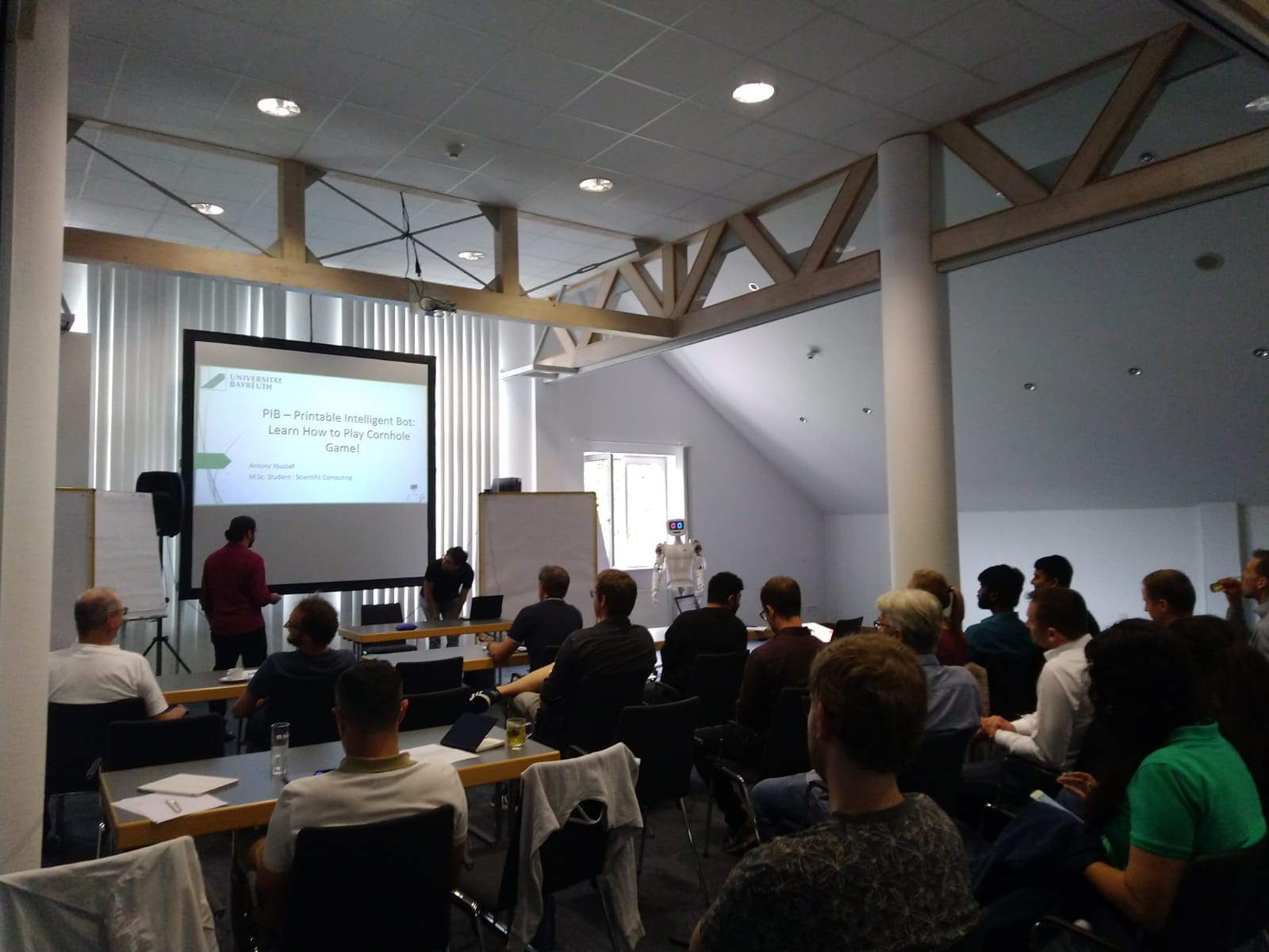 The picture shows students giving a presentation in front of an audience, with the robot pib in the background.