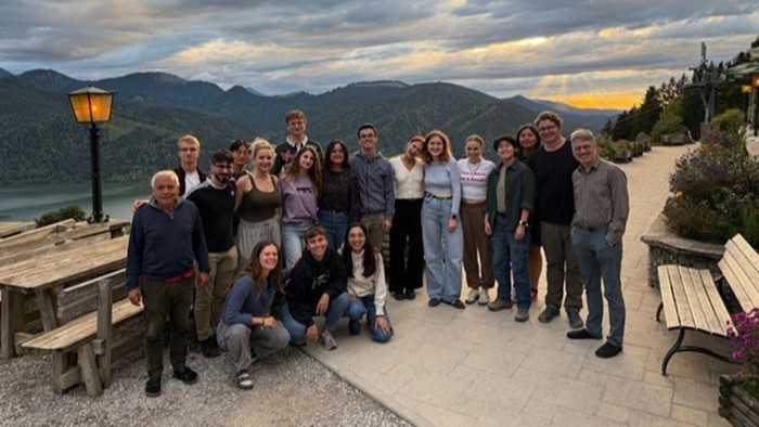 A group of young people is standing together outdoors in front of an impressive mountain and lake landscape.