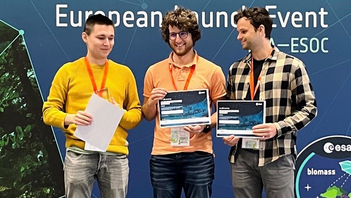 Das Bild zeigt drei junge Männer, die auf einer Bühne vor einem Banner mit der Aufschrift „European Launch Event“ stehen. Zwei von ihnen halten Zertifikate in den Händen, alle drei wirken fröhlich und sind mit Namensschildern ausgestattet.