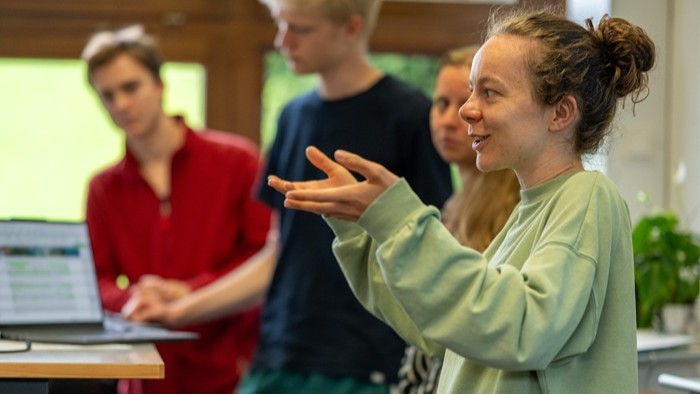 A person is standing in a room gesturing while others in the background listen near a laptop.
