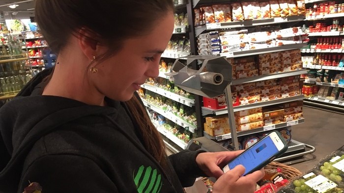A young women is standing in the supermarkt holding her mobilphone where the app is displayed.