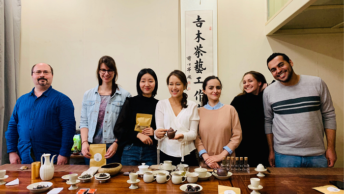 Seven persons stand behind a wooden table full of traditional tea utensils.