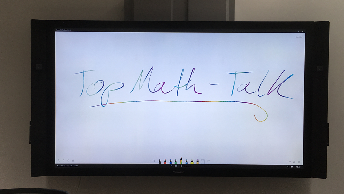 Auf einem Whiteboard steht in bunter Schrift der Veranstaltungstitel, „TopMath-Talk“, davor sind die Lehnen von drei leeren Stühlen zu sehen.